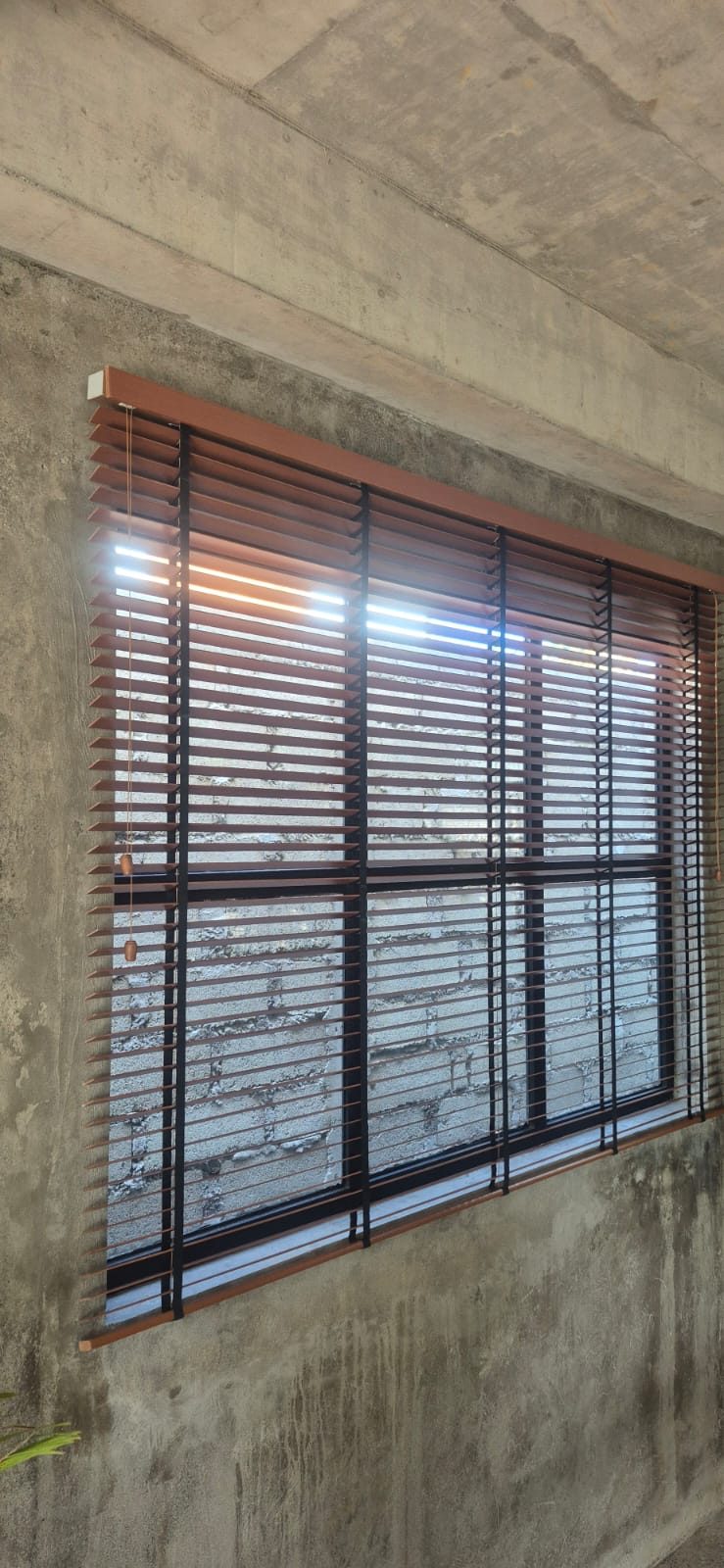 Pemasangan wooden blind (tirai kayu) warna cokelat hangat yang mengoptimalkan kontrol pencahayaan alami, sirkulasi udara, dan estetika bernuansa natural pada jendela ruangan.